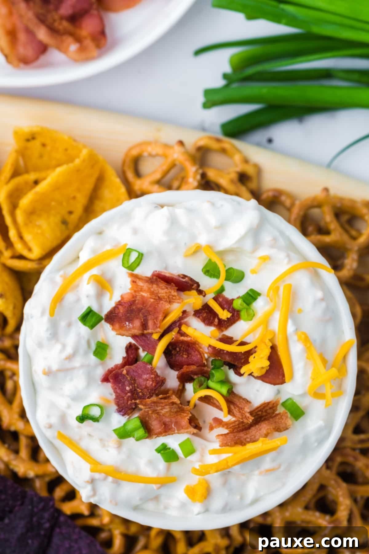 Deconstructed Baked Potato Dip 6 An overhead shot of a beautifully garnished bowl of Loaded Baked Potato Dip, topped with green onion, shredded cheddar cheese, and crumbled bacon, surrounded by corn chips and pretzels for dipping.