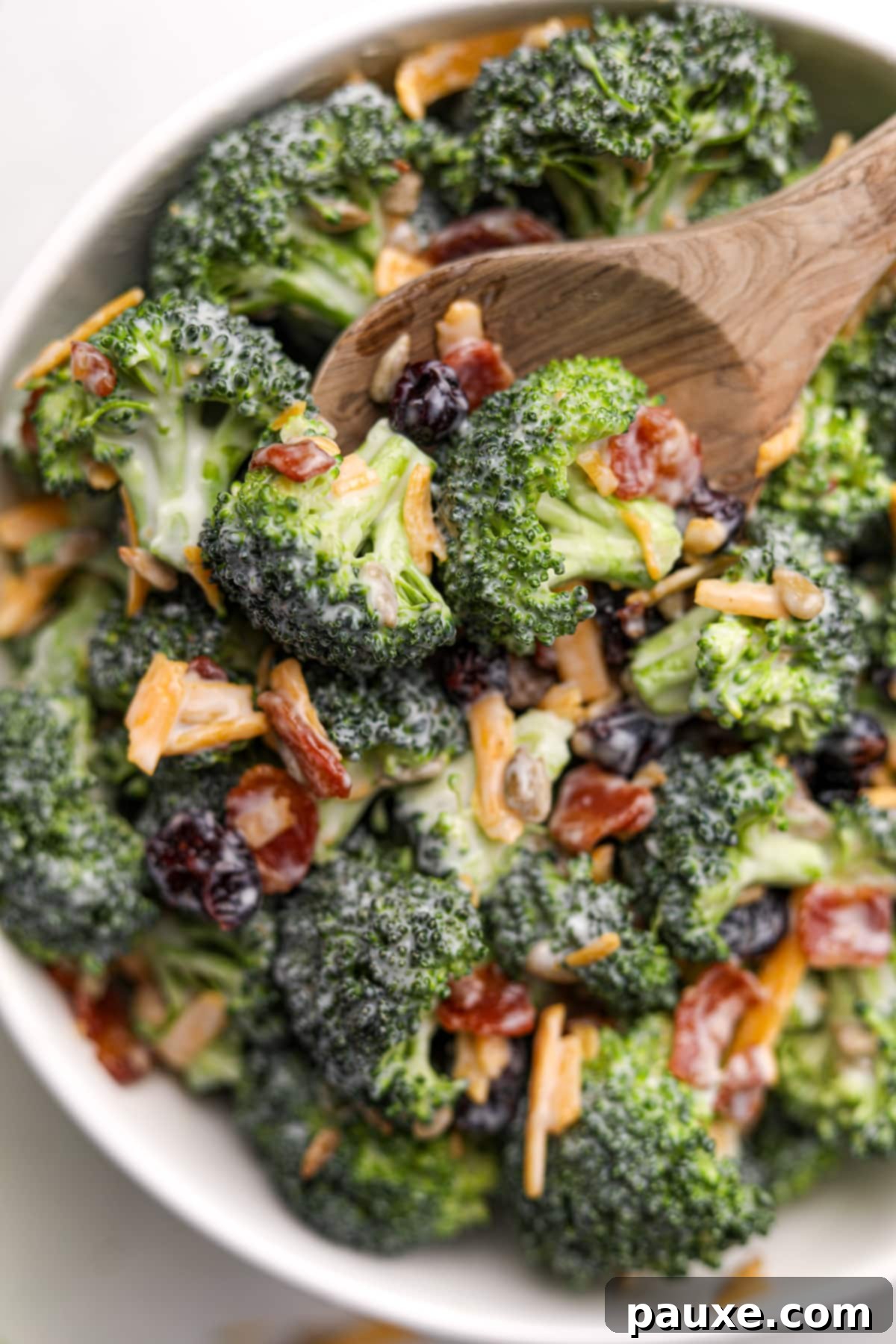 Creamy Crunch Broccoli Salad 2 A wooden spoon taking a small portion from a large bowl of creamy broccoli salad, garnished with crispy bacon bits and shredded cheddar cheese.