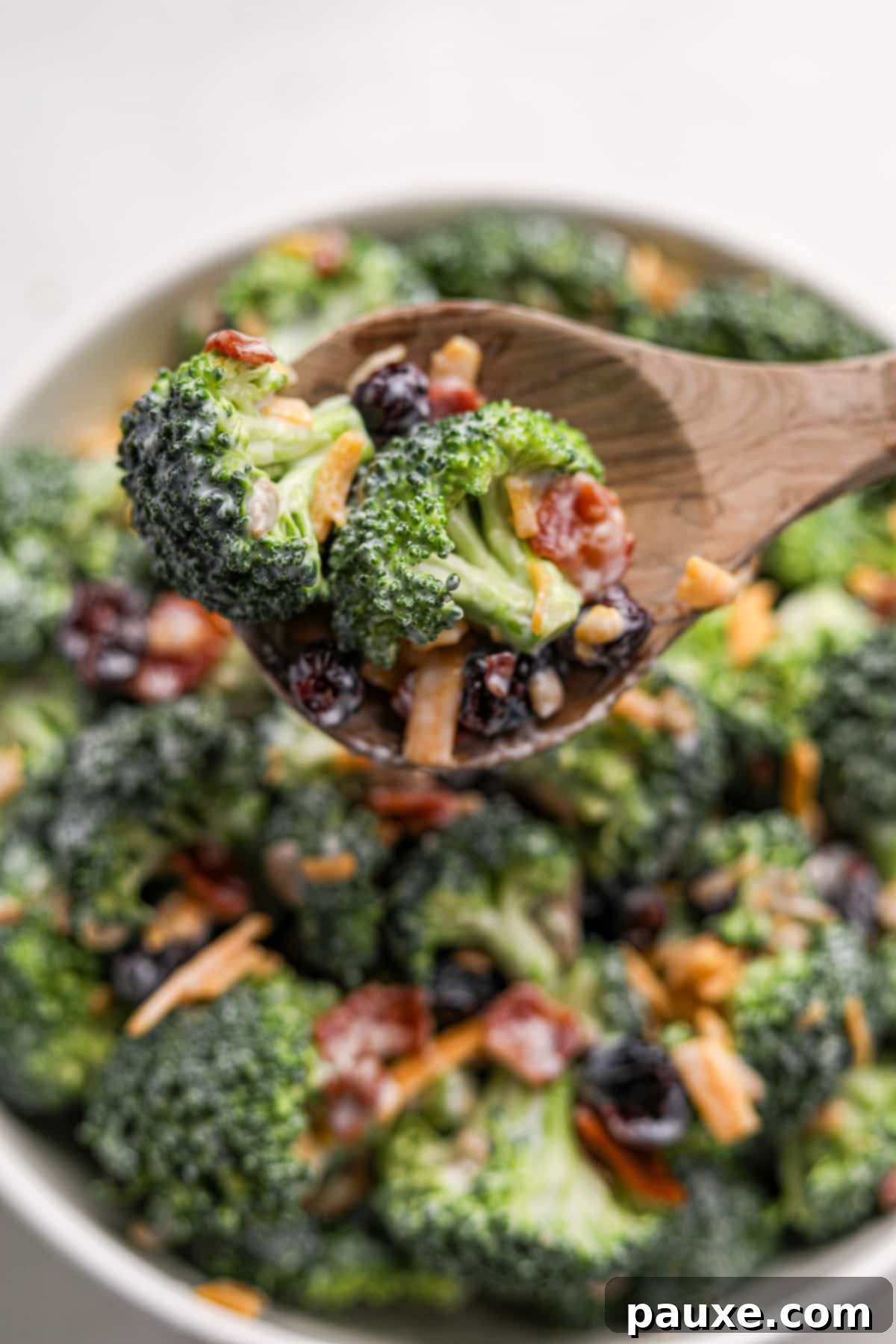Creamy Crunch Broccoli Salad 14 A wooden spoon scooping a generous portion of creamy broccoli salad from a large white bowl, featuring raw broccoli, crispy bacon, and sharp cheddar cheese.