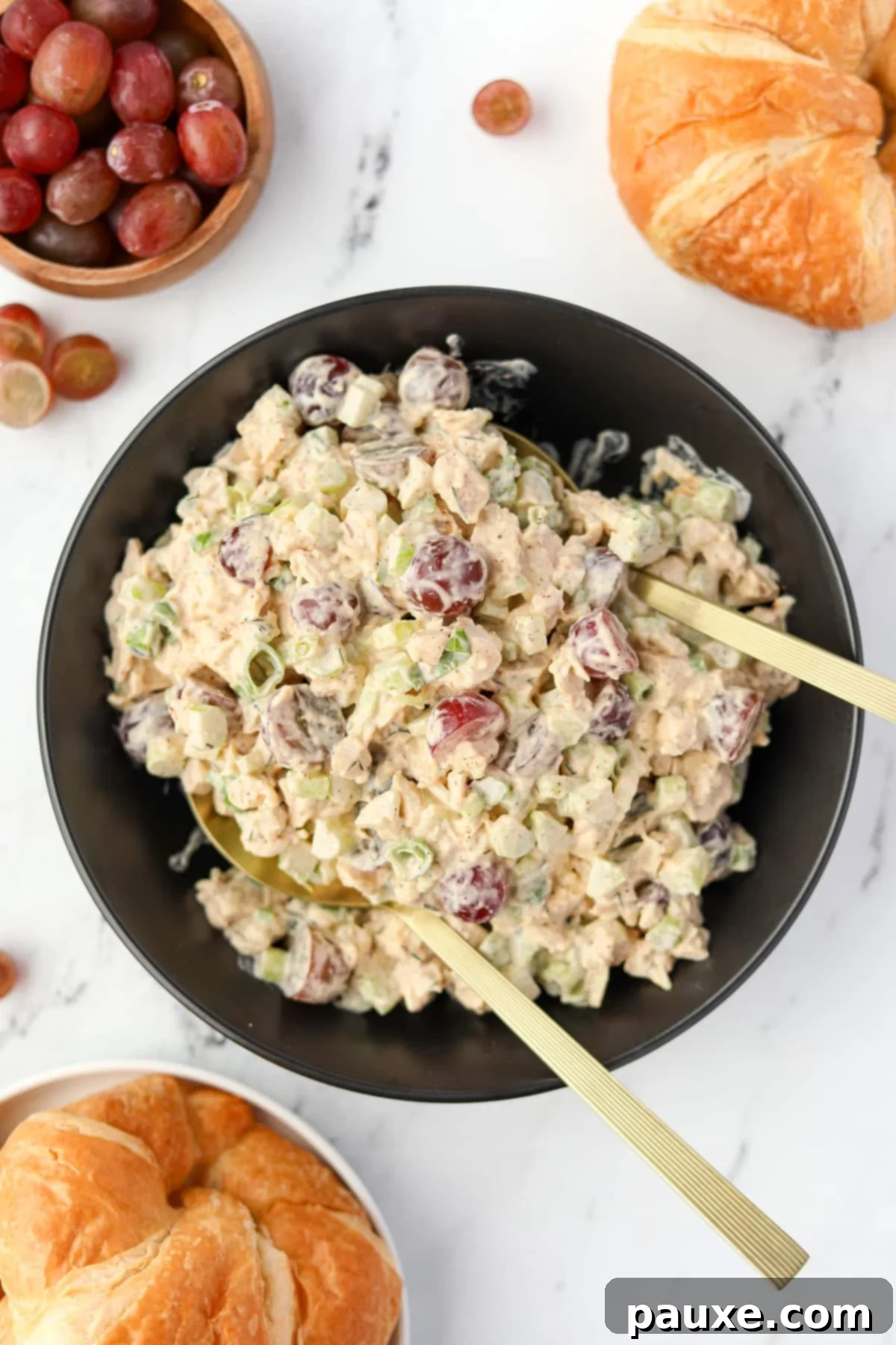 A bowl of creamy chicken salad with grapes, celery, and green onions being tossed with serving spoons.