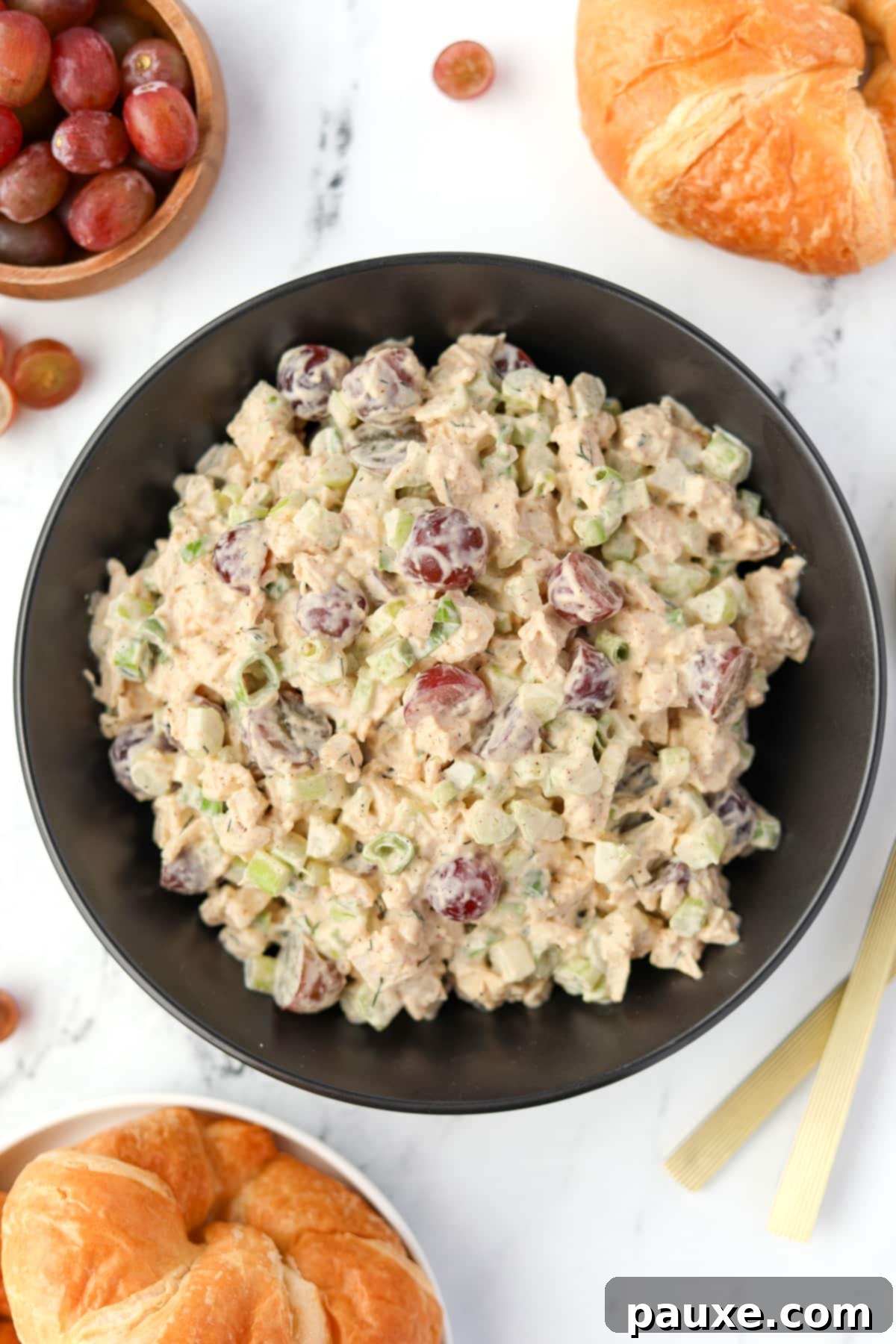 A beautifully presented black bowl of chicken salad with grapes, accompanied by fresh grapes and warm croissants.