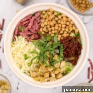 A bowl of chopped salad, featuring mozzarella cheese, artichokes, sun dried tomatoes, chickpeas, salami, and basil.