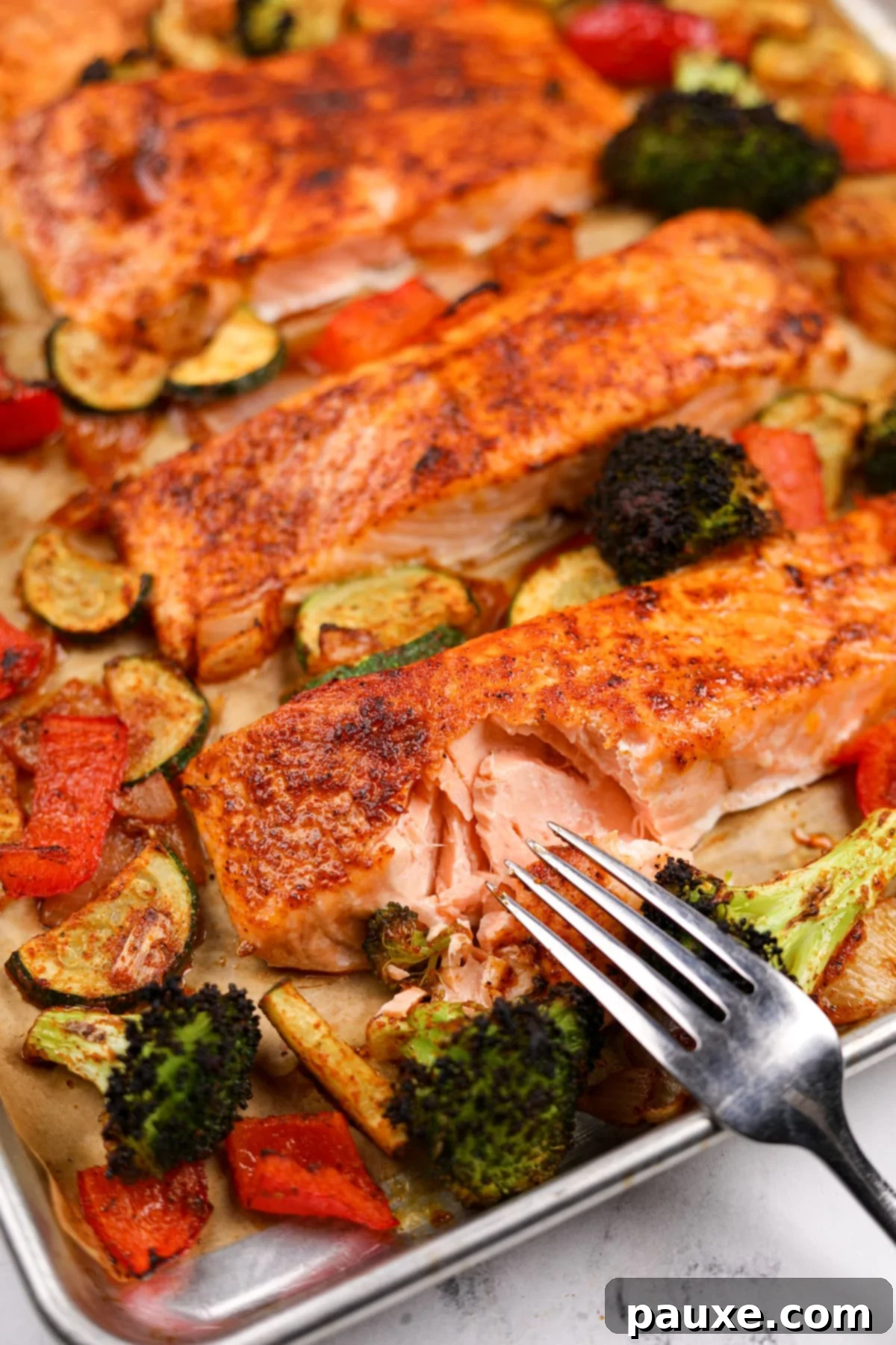 A fork flaking a piece of salmon on a pan, surrounded by roasted vegetables.
