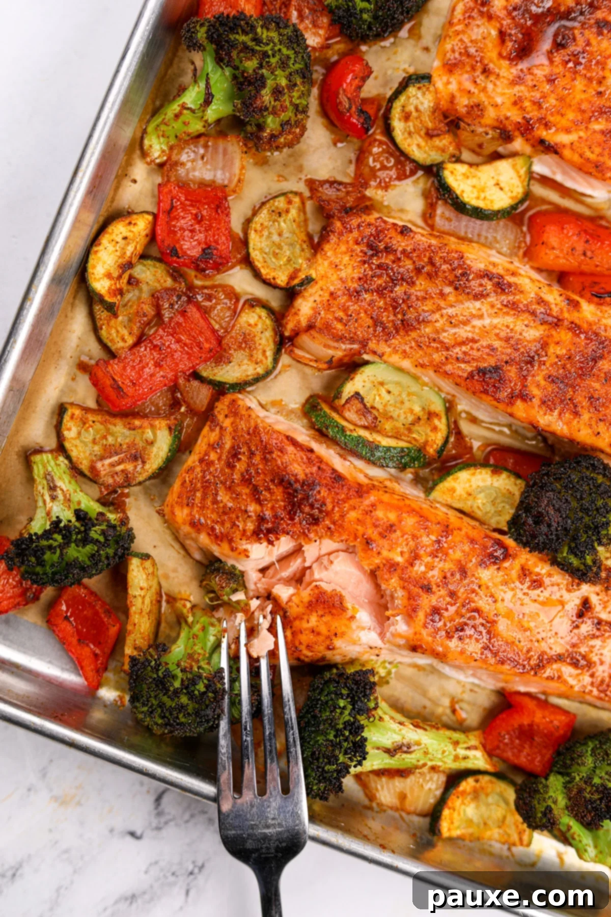 A fork flaking a fillet of salmon, surrounded by various vegetables including broccoli, bell pepper, and zucchini.