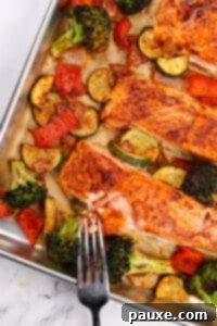 A fork flaking a fillet of salmon, surrounded by various vegetables including broccoli, bell pepper, and zucchini.