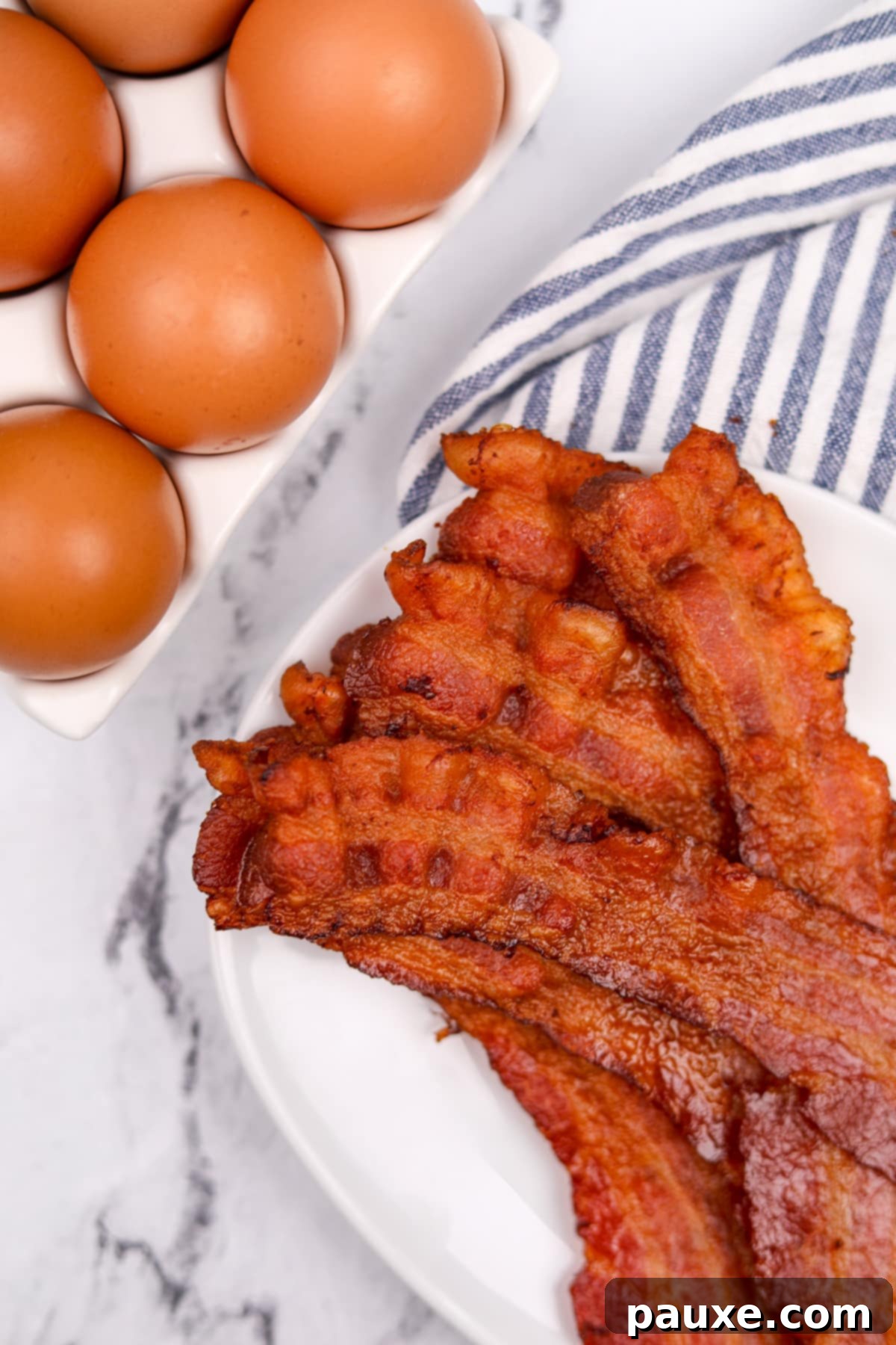 Perfectly Crispy Oven Bacon 7 Crispy bacon slices on a white plate, with blurred scrambled eggs in the background, making a delicious breakfast scene.