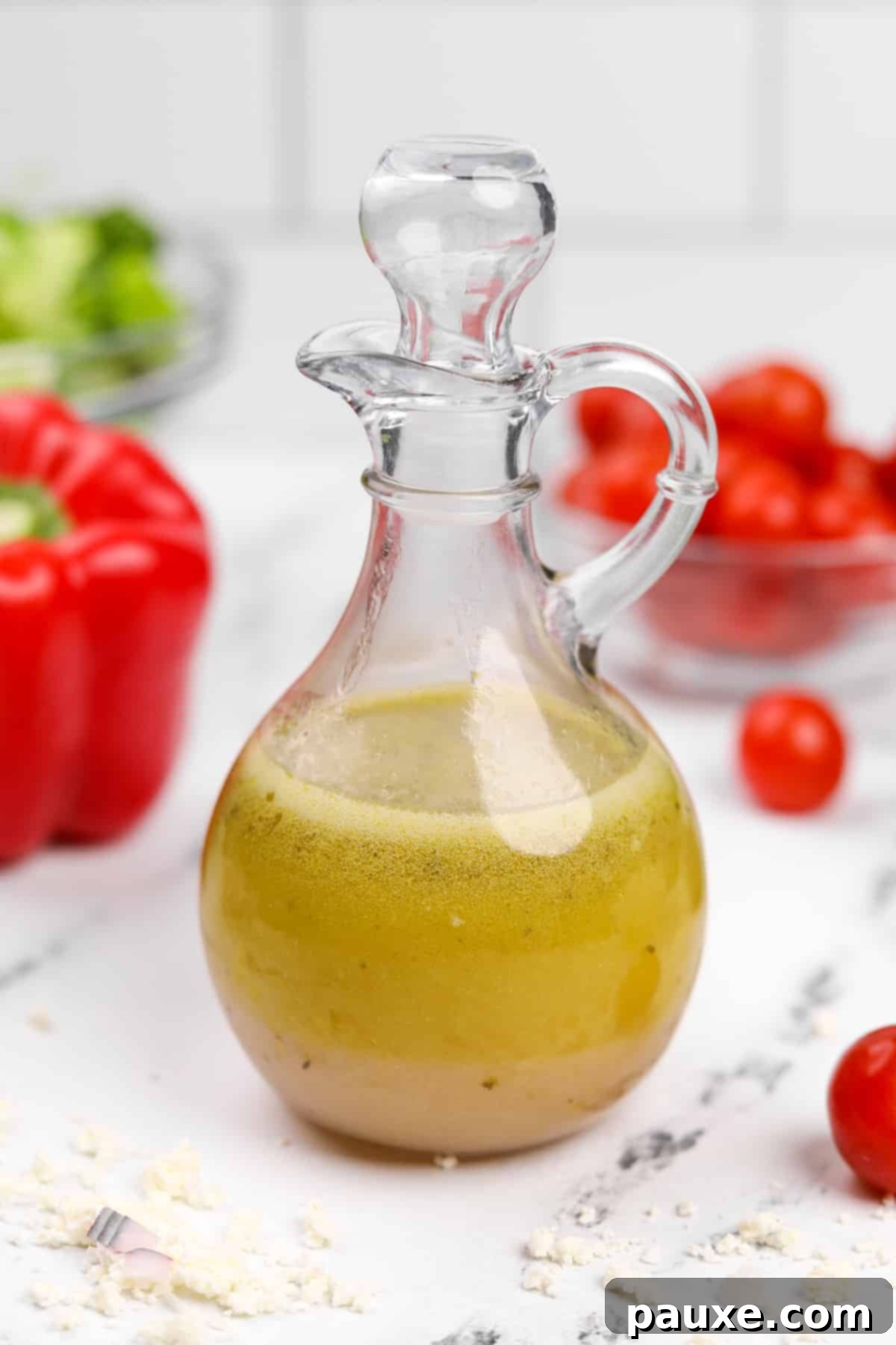 Zesty Mediterranean Vinaigrette 2 A cruet filled with golden homemade Greek salad dressing, surrounded by fresh ingredients like red pepper, tomatoes, and herbs.