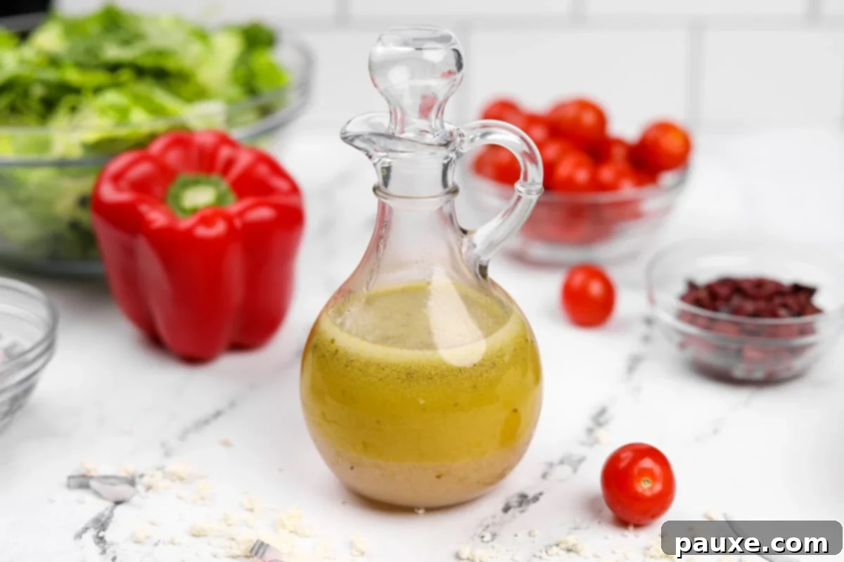Zesty Mediterranean Vinaigrette 7 A clear glass cruet holding homemade Greek salad dressing, with a background blur of fresh salad ingredients like red pepper, lettuce, and tomatoes.