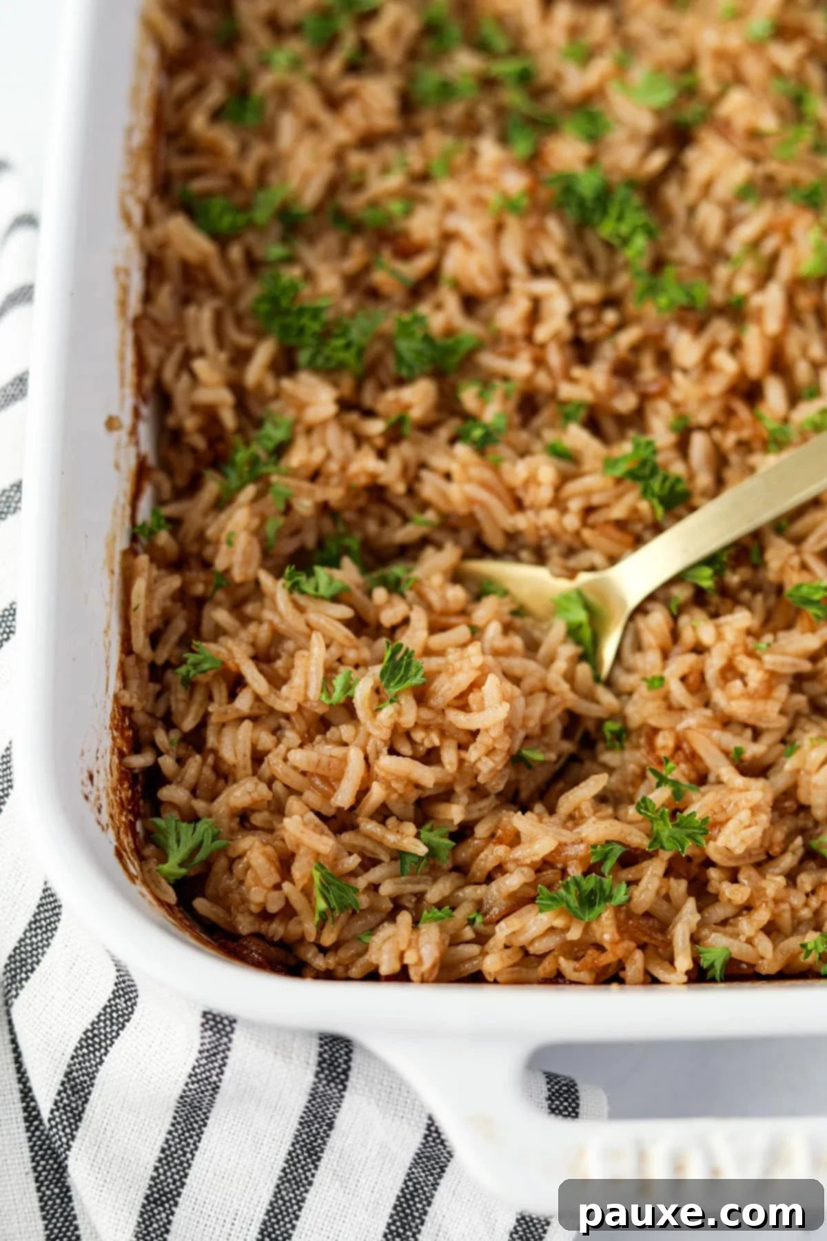Rich Beef Consommé Rice 5 A close-up shot of a serving spoon gracefully scooping a pile of perfectly fluffy, golden-baked rice from a white baking dish, showcasing its inviting texture.