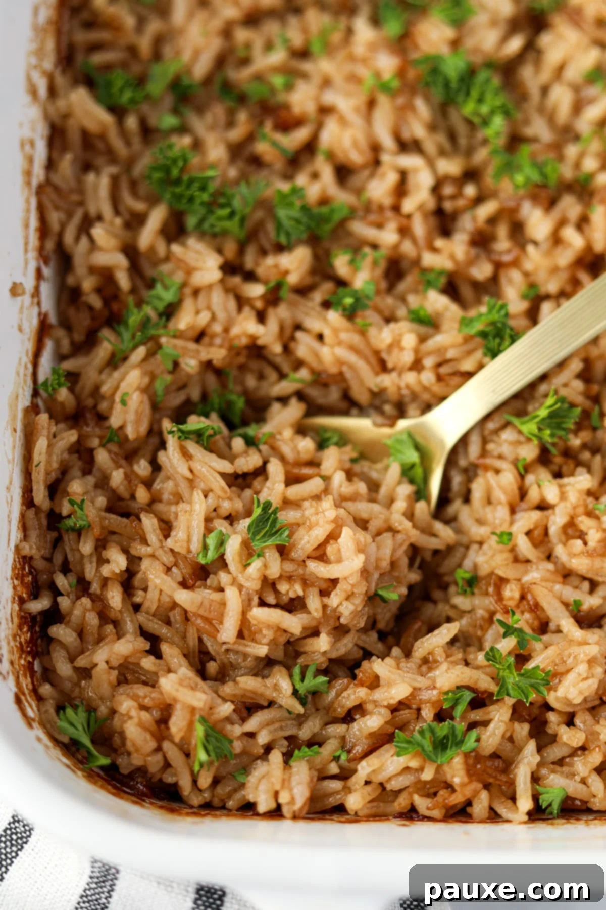 Rich Beef Consommé Rice 7 A close-up of a serving spoon scooping a generous portion of fluffy, golden-baked rice from a white baking dish, showing its appealing texture.