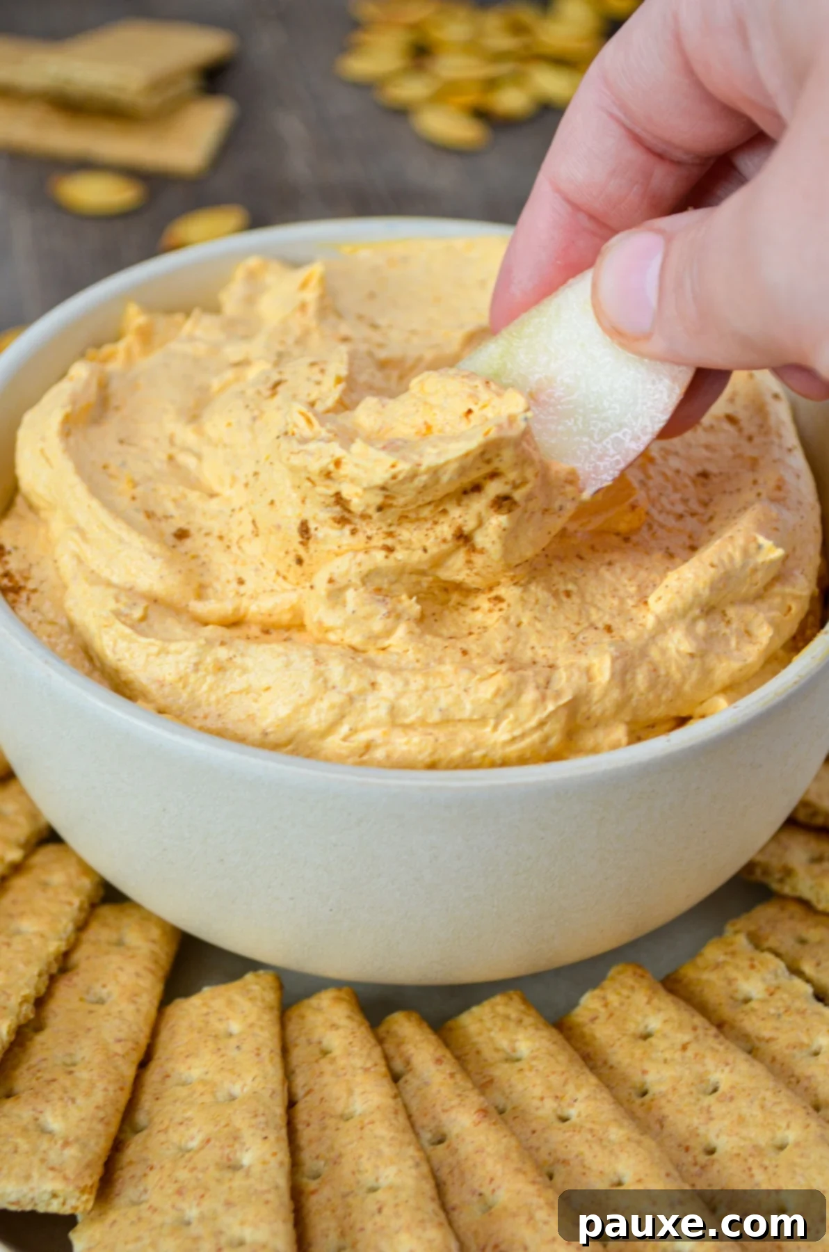 A bowl of pumpkin fluff dip, surrounded by graham crackers. A hand dips an apple slice into the orange colored dip.