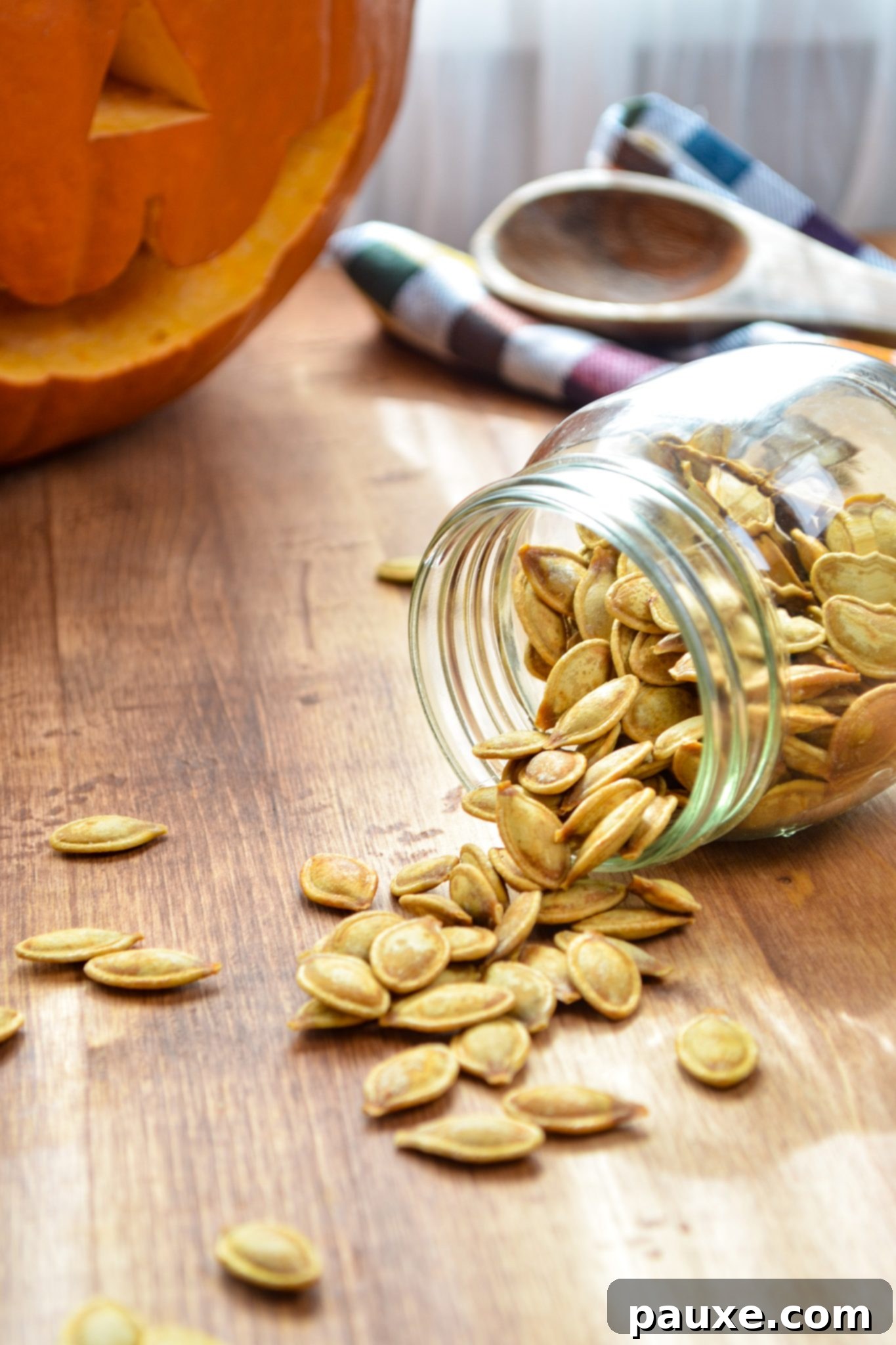 Crispy Air Fryer Pumpkin Seeds 11 An artfully arranged scene of roasted pumpkin seeds spilling from a glass jar onto a wooden table, with a festive, carved pumpkin providing a warm, autumnal backdrop.