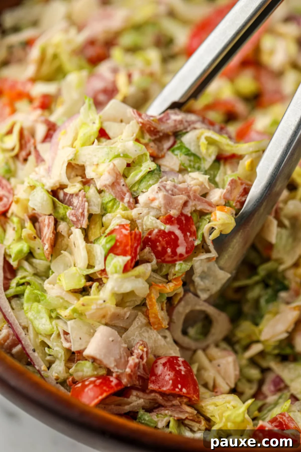 Hero Salad Bowl 2 Tongs taking a portion of chopped Grinder Salad from a large wooden bowl, showcasing crisp lettuce, deli meats, and creamy dressing.