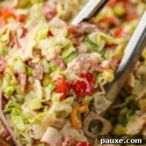 Hero Salad Bowl 9 A pair of tongs picking up a portion of chopped Italian Grinder Salad from a wooden bowl, showing the creamy dressing and colorful ingredients.