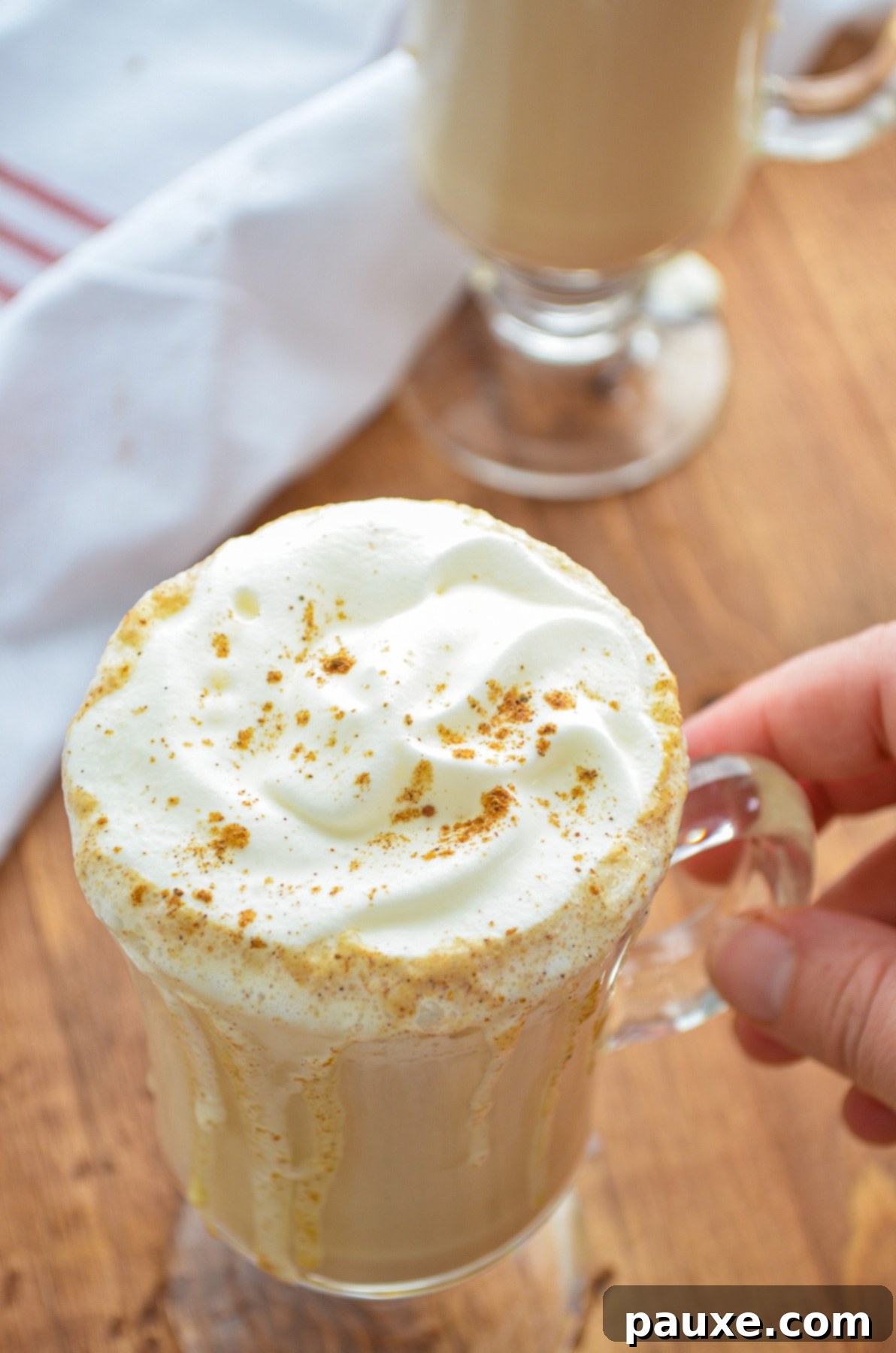 Homemade Instant Pot Pumpkin Spice Latte 6 A hand gracefully lifting a clear glass mug, showcasing a rich, layered pumpkin spice latte topped with a luxurious swirl of whipped cream, in a cozy indoor setting.