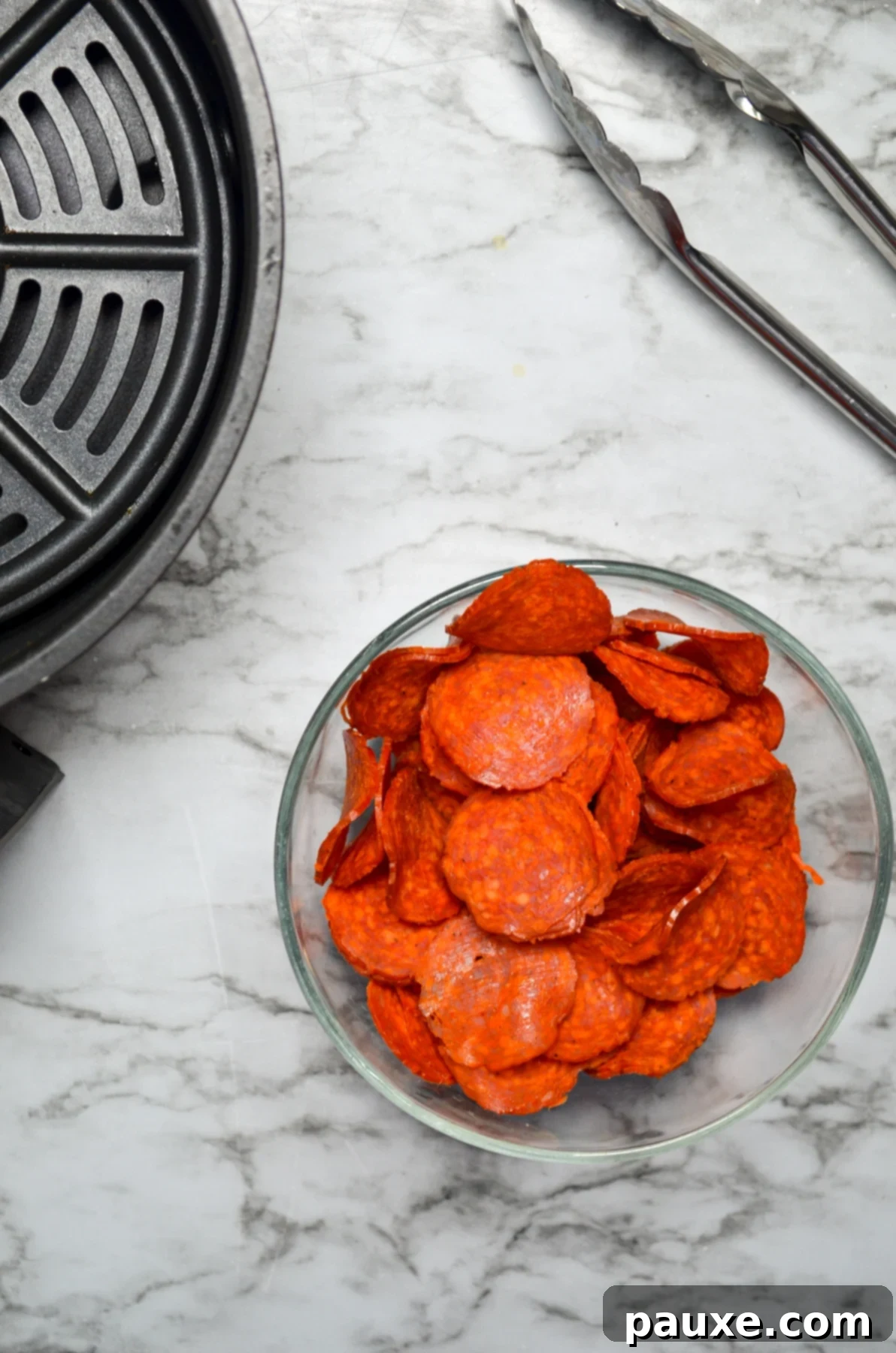 Air Fried Crispy Pepperoni 4 A bowl of pepperoni, with an air fryer basket and a pair of tongs on the side.