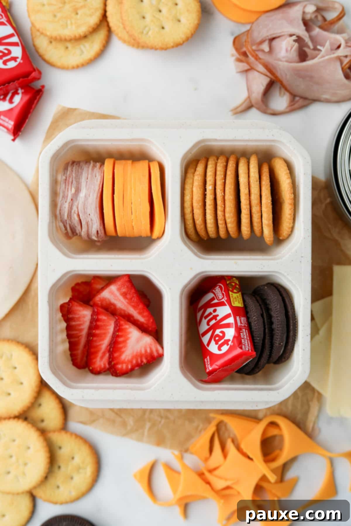 Craft-Your-Own Lunch Kits 6 A perfectly arranged lunch container, featuring neatly stacked cheese and crackers, fresh fruit, a mini chocolate bar, and small cookies, ready for a delicious meal.