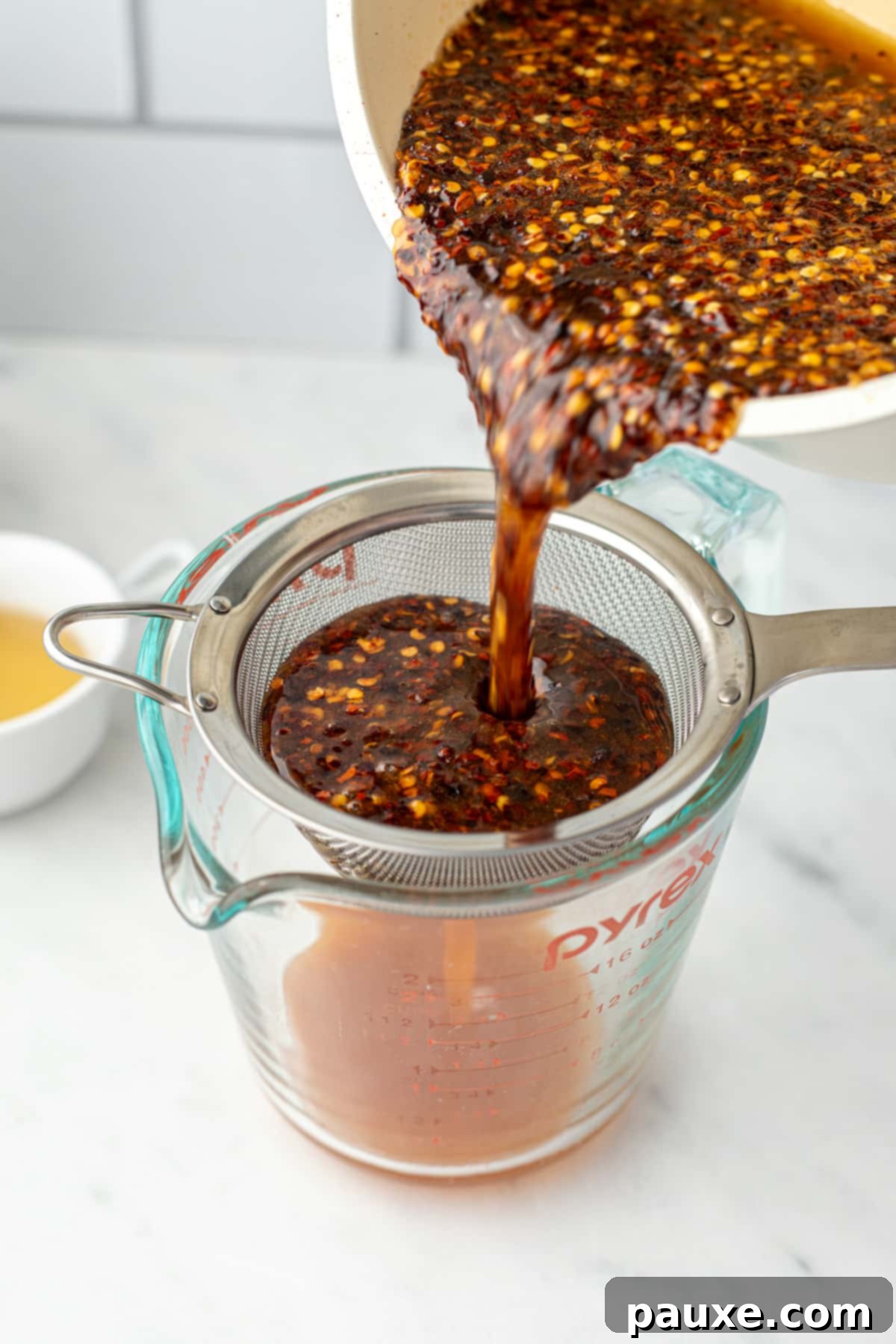 Crafting Homemade Hot Honey 6 Pouring infused hot honey through a fine mesh strainer into a glass measuring cup, separating the chili flakes.