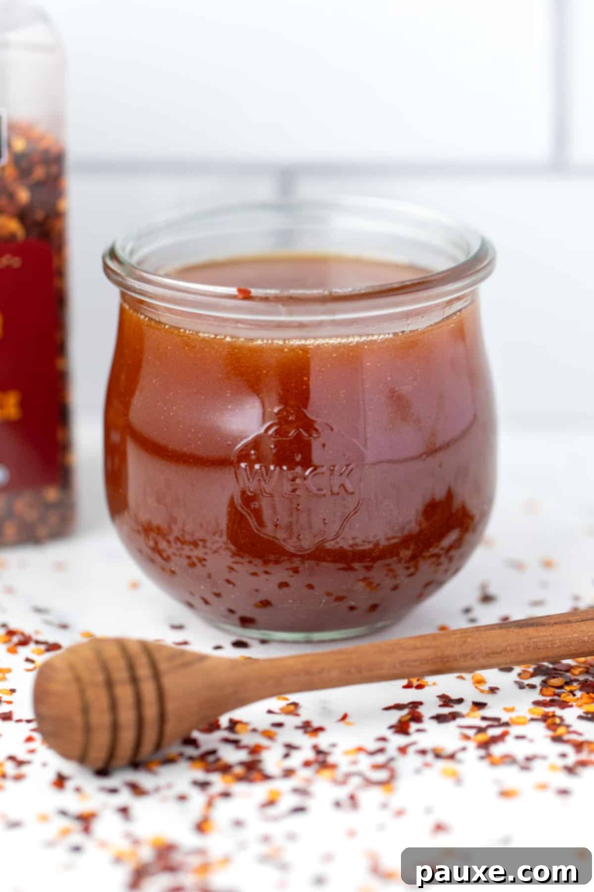 Crafting Homemade Hot Honey 8 A weck jar filled with hot honey, with a honey dipper on the counter next to scattered crushed red pepper flakes, showcasing the finished product.