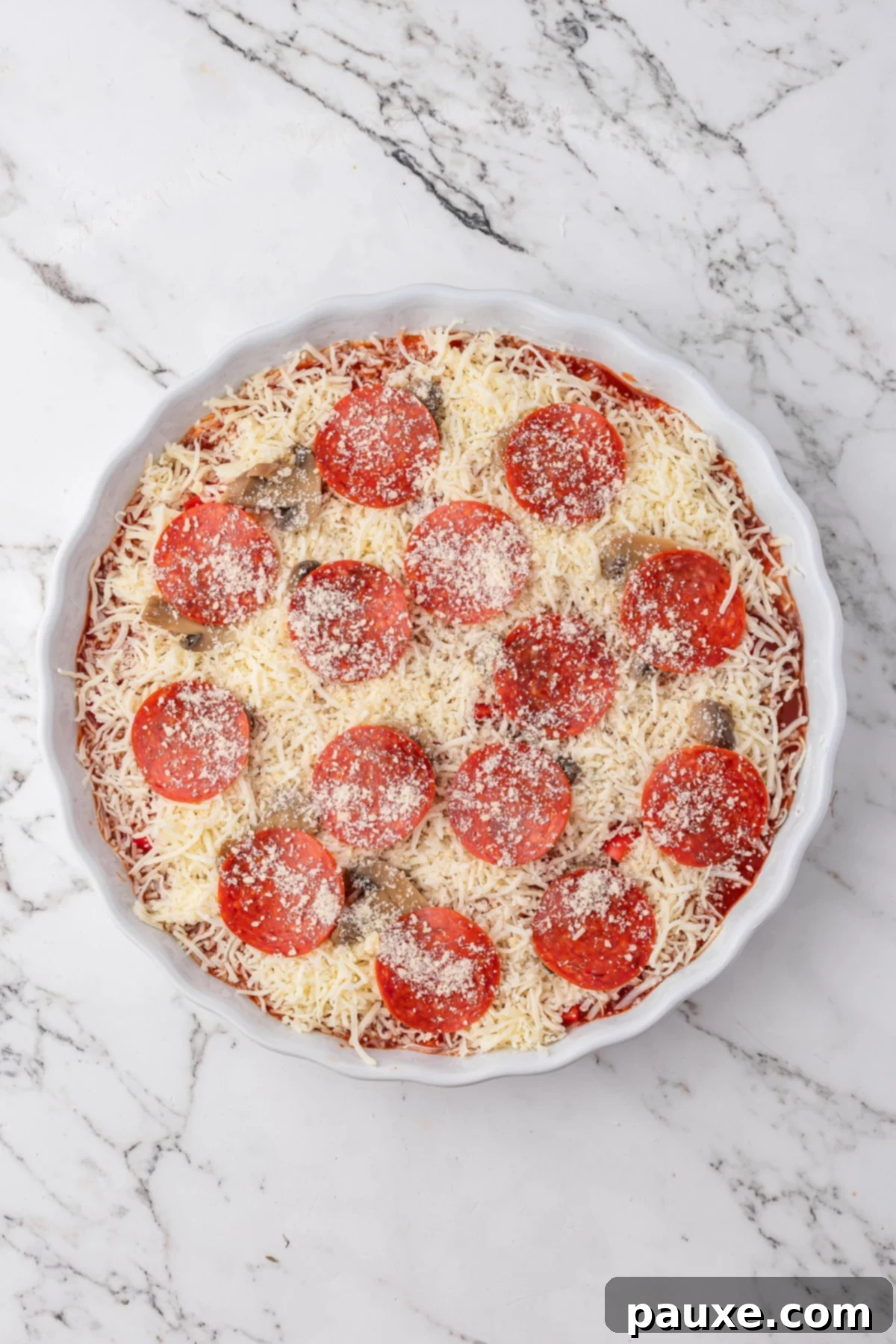 Baked Pepperoni Pizza Dip 7 A round baking dish with the layered pizza dip, generously topped with shredded mozzarella cheese, pepperoni slices, and a sprinkle of Parmesan cheese, ready for the oven.