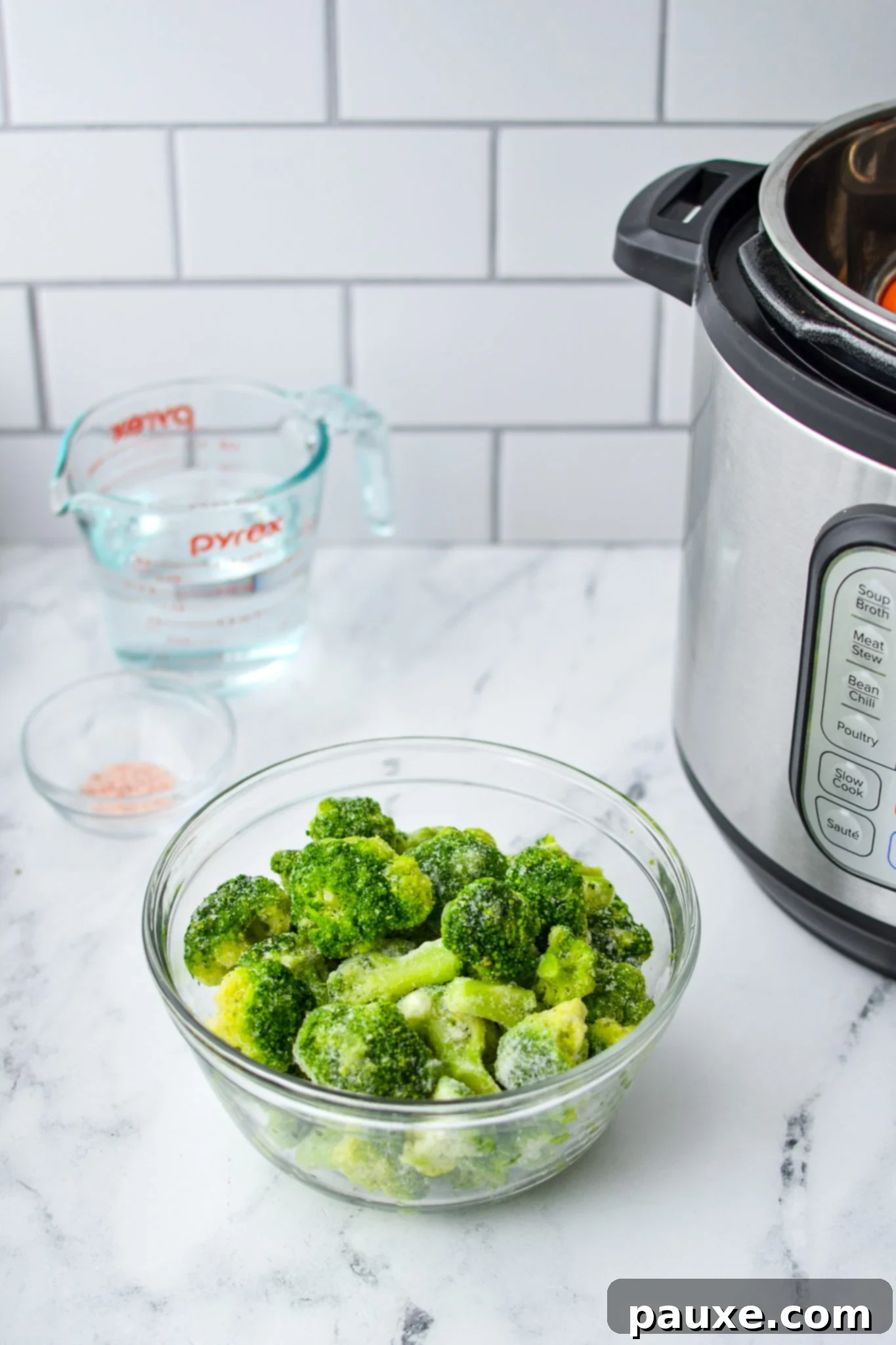 Perfect Instant Pot Frozen Broccoli 4 A bowl of frozen broccoli on a white marble countertop. A measuring cup of water, some salt, and an Instant Pot in the background, ready for cooking.
