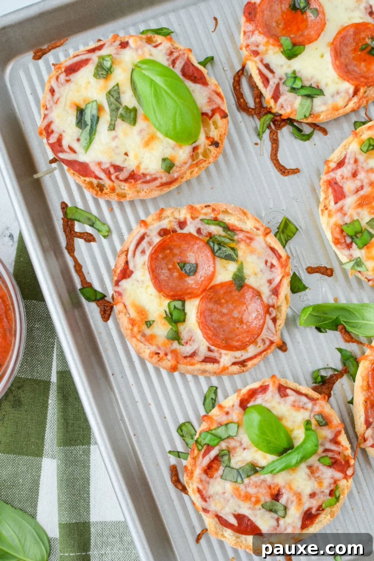 A baking sheet with English muffin pizzas topped with pepperoni and basil.