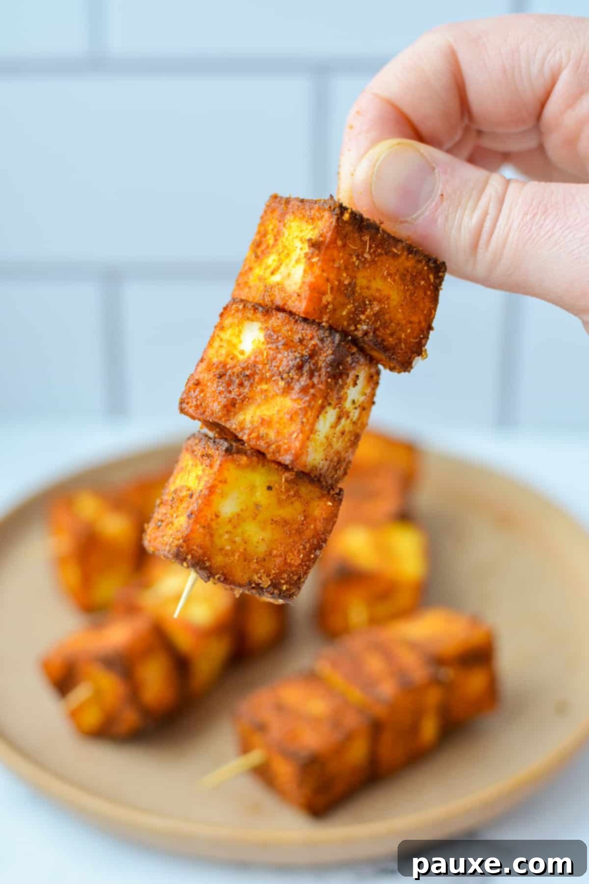 A hand holding a skewer with three cubes of seasoned paneer speared onto it, highlighting the vibrant color and texture.
