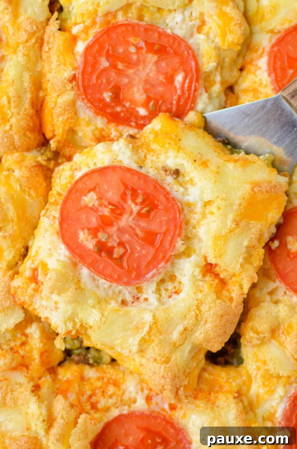 The Duke's Hearty Casserole 3 A spatula serving a portion of cheesy John Wayne Casserole, topped with fresh tomato slices, from a hot baking dish, ideal for a hearty dinner.