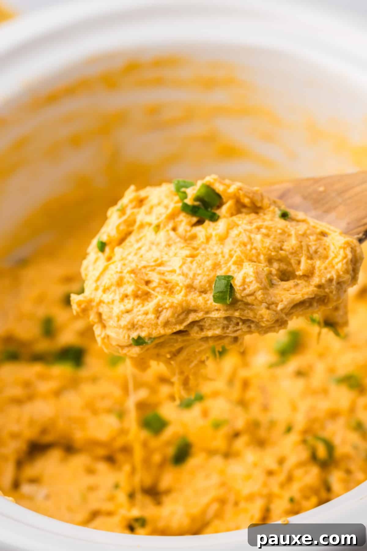 Zesty Slow Cooker Buffalo Chicken Dip 14 A wooden spoon carefully scooping a generous portion of hot and cheesy buffalo chicken dip from a white slow cooker.