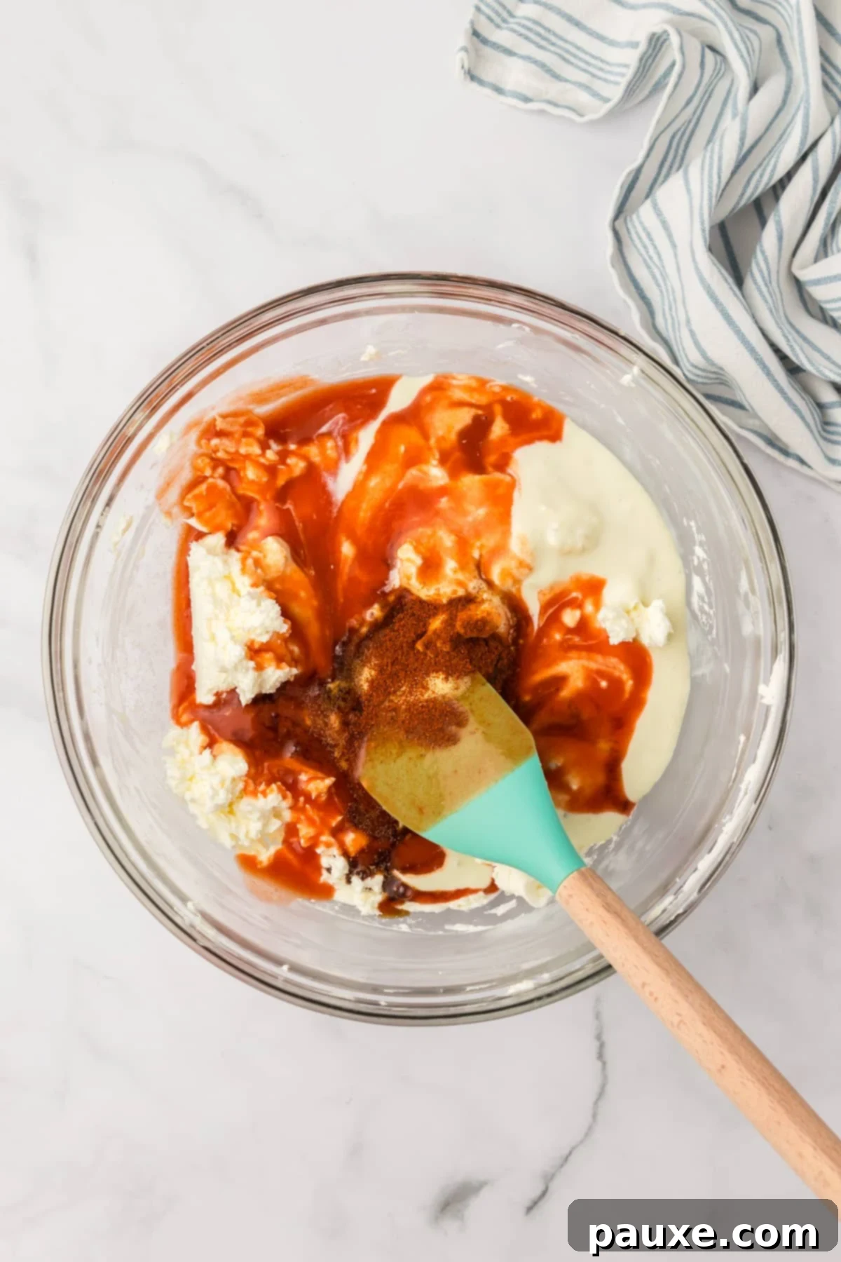 Zesty Slow Cooker Buffalo Chicken Dip 5 A glass bowl with a smooth cream cheese mixture, with a stream of vibrant cayenne pepper sauce being added.