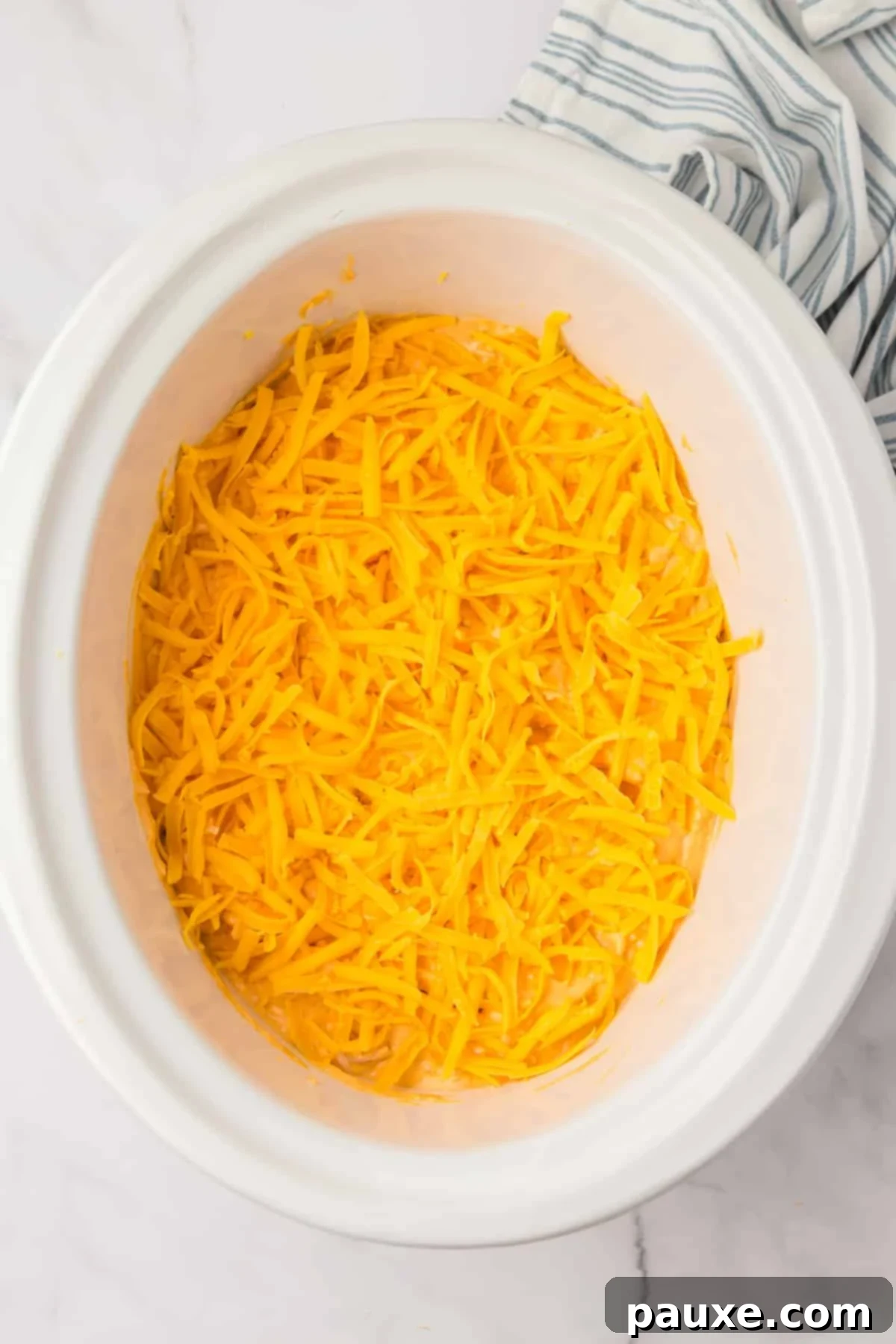 Zesty Slow Cooker Buffalo Chicken Dip 9 A white slow cooker with the cheesy dip inside, now beautifully topped with an extra layer of shredded cheddar cheese, ready for cooking.