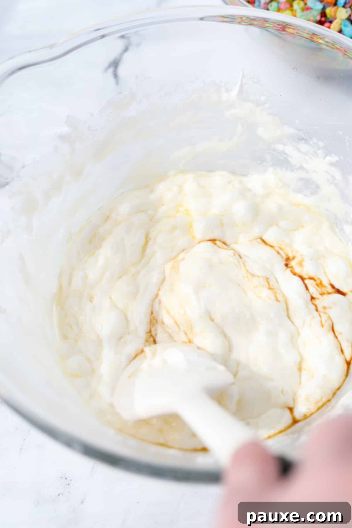 Colorful Pebbles Bars 5 A hand stirring melted marshmallows with butter and vanilla extract in a bowl, creating a smooth, glossy mixture.