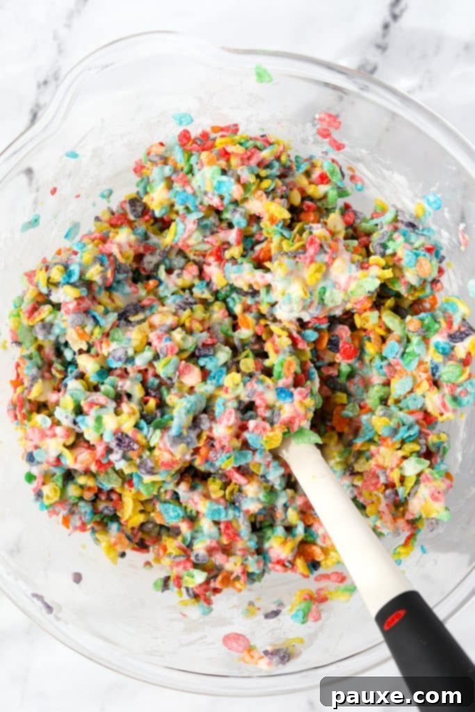 Colorful Pebbles Bars 6 A large bowl filled with vibrant Fruity Pebbles cereal being gently folded into the sticky marshmallow mixture with a silicone spatula.