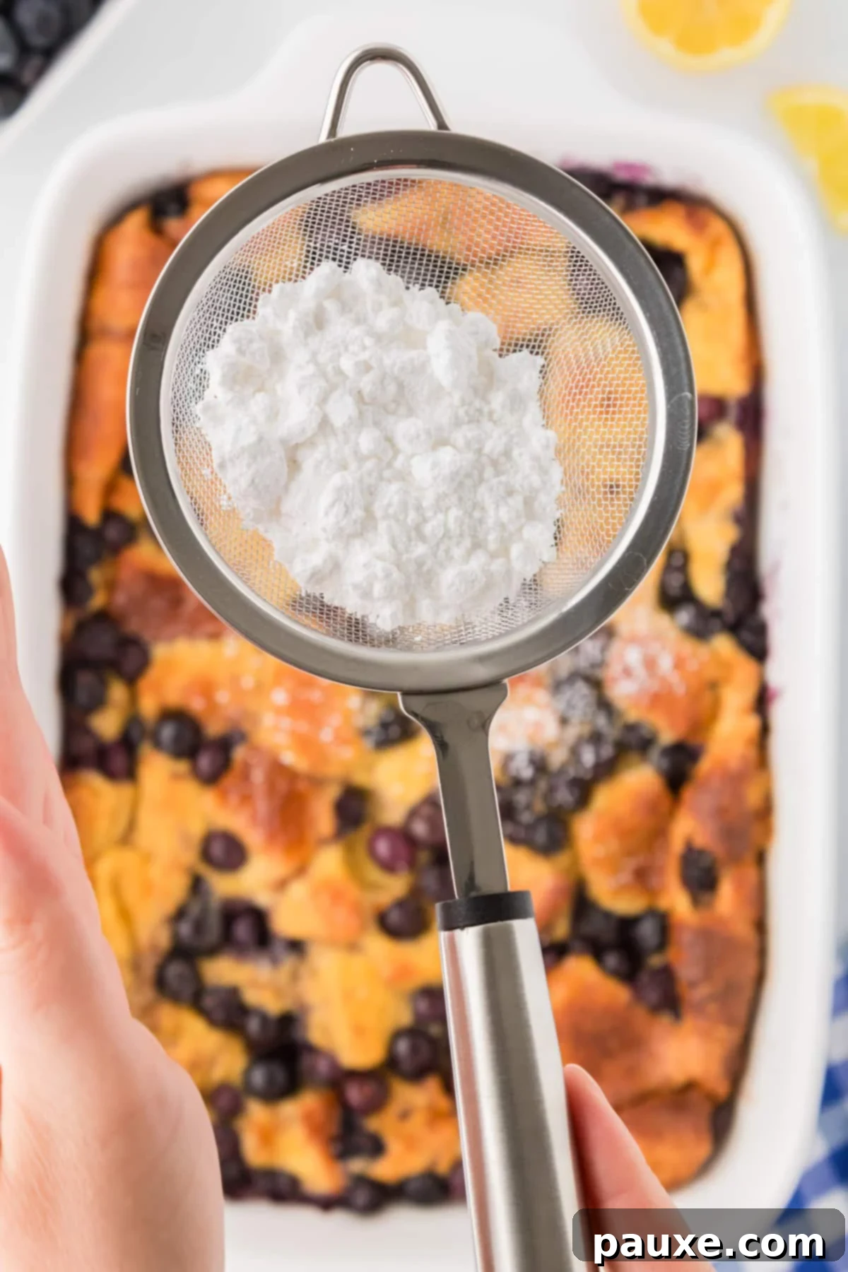 Overnight Brioche French Toast Bake 11 A hand using a small sifter to dust powdered sugar over the warm, golden-brown surface of the French toast casserole, adding a finishing touch.