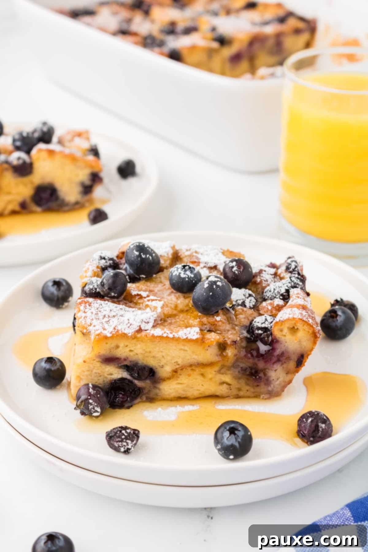 Overnight Brioche French Toast Bake 13 A perfectly baked square of brioche French toast casserole, artfully garnished with fresh blueberries and a generous drizzle of golden maple syrup, on a white plate.