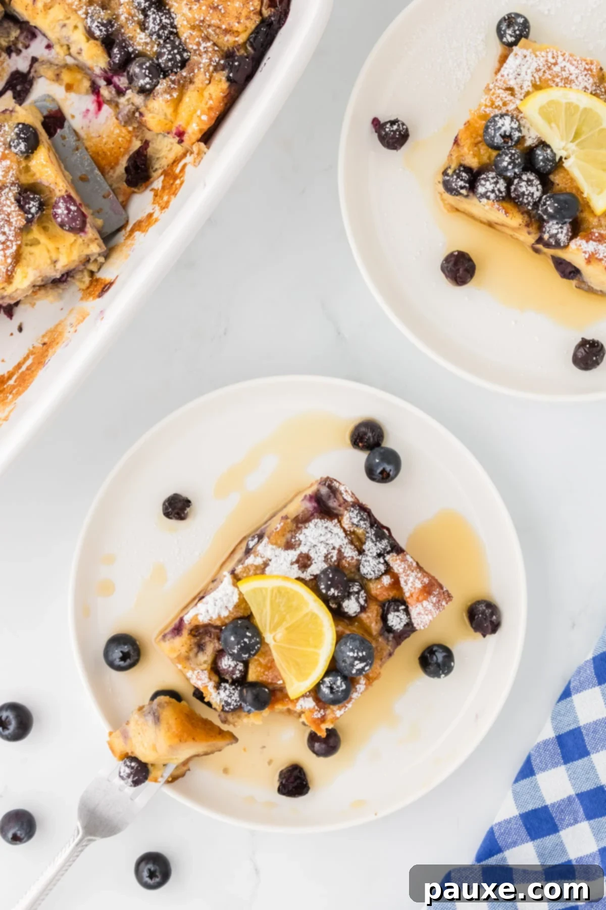 Overnight Brioche French Toast Bake 15 A serving of baked brioche French toast casserole on a white plate, beautifully garnished with lemon slices, fresh blueberries, and a drizzle of maple syrup, against a rustic wooden background.