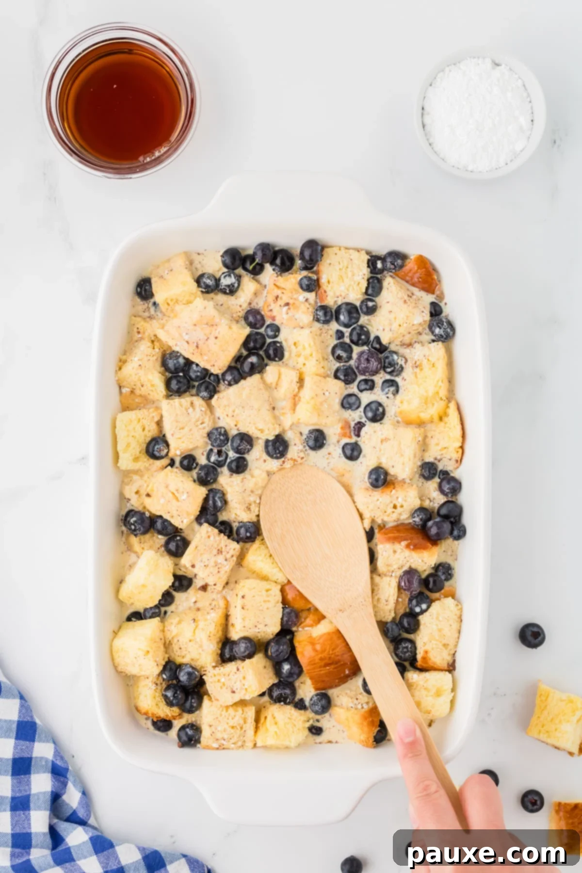 Overnight Brioche French Toast Bake 8 A wooden spoon gently pressing down on brioche bread cubes soaking in a creamy custard mixture within a baking dish, ensuring all pieces are fully submerged.