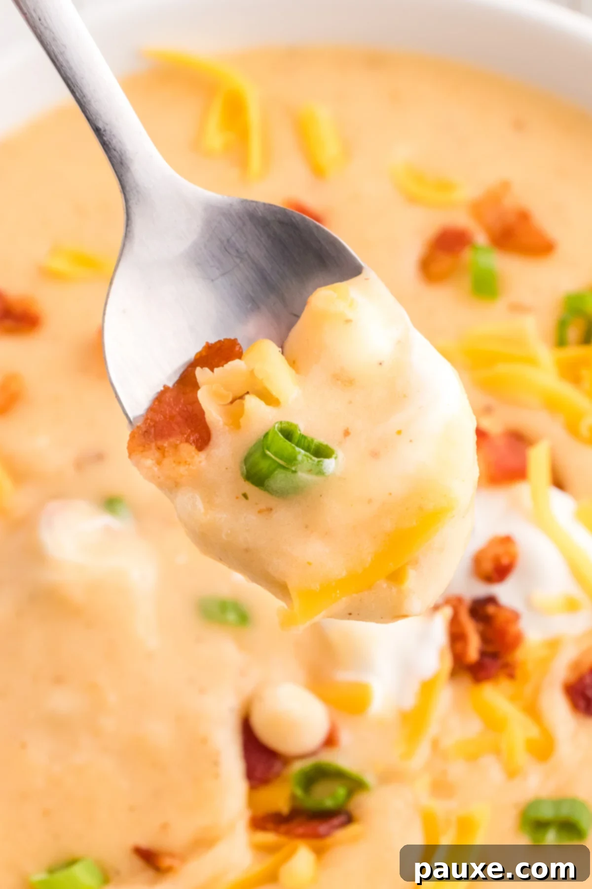 A close-up of a spoon laden with cheesy potato soup, garnished with crispy bacon and fresh green onions.