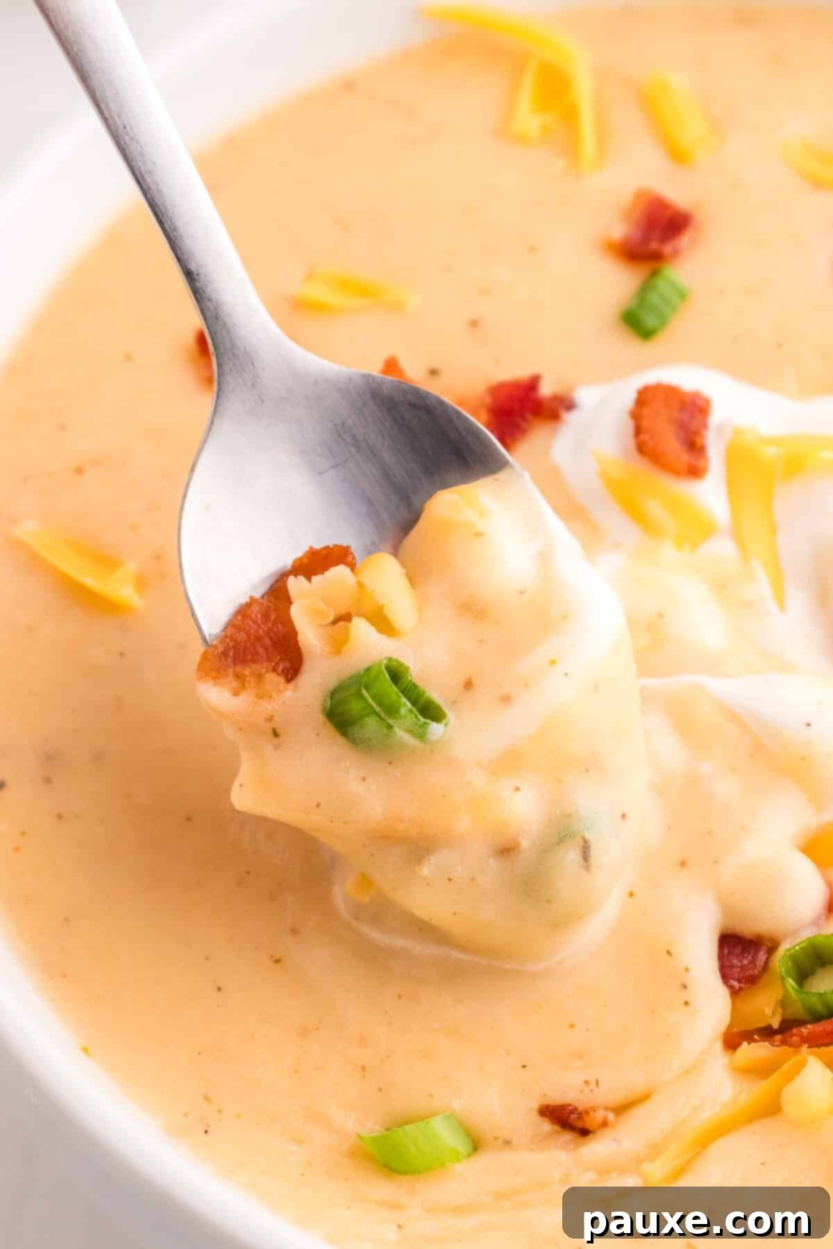 A spoon taking a generous portion of cheesy baked potato soup, adorned with fresh green onions and crispy bacon bits.
