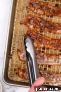 Decadent Glazed Bacon 16 A pair of tongs flipping bacon on a baking sheet.