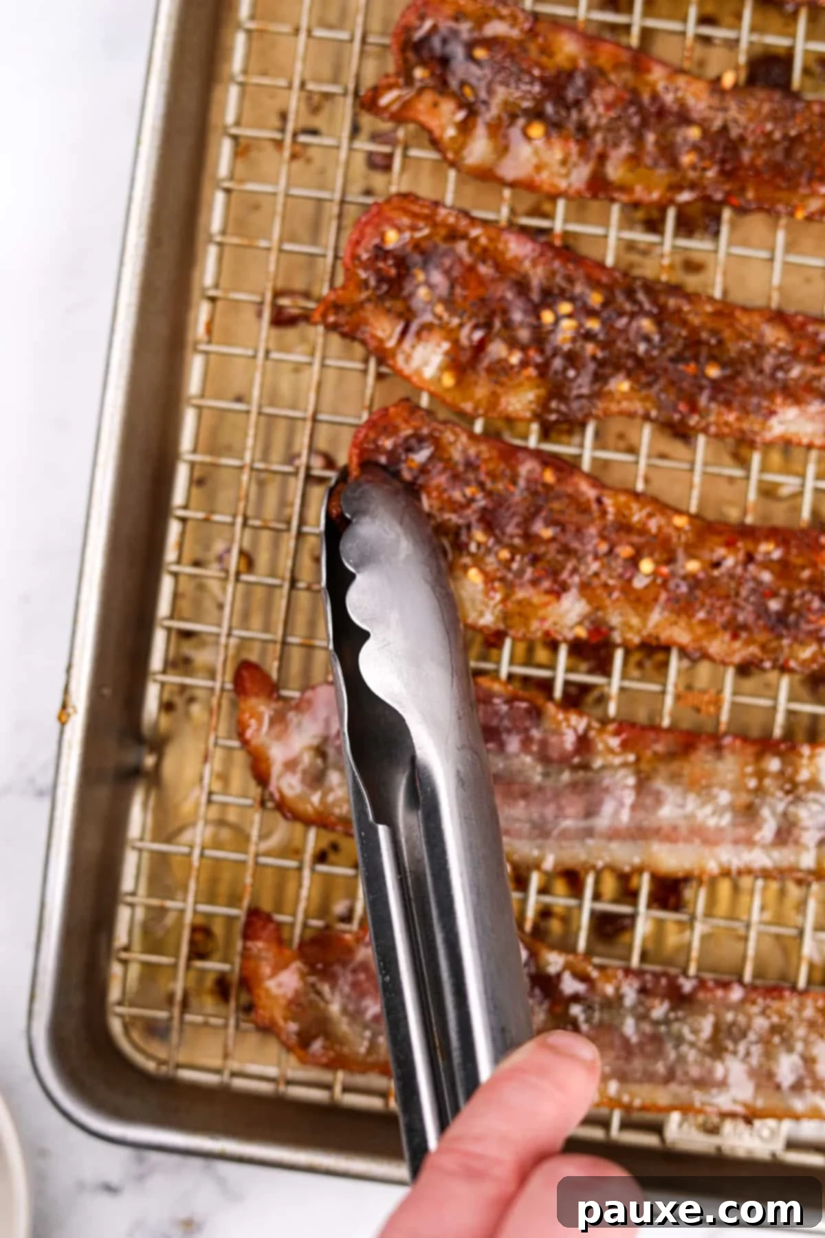 Decadent Glazed Bacon 8 A pair of tongs carefully flipping individual bacon slices on a baking sheet to coat the other side.