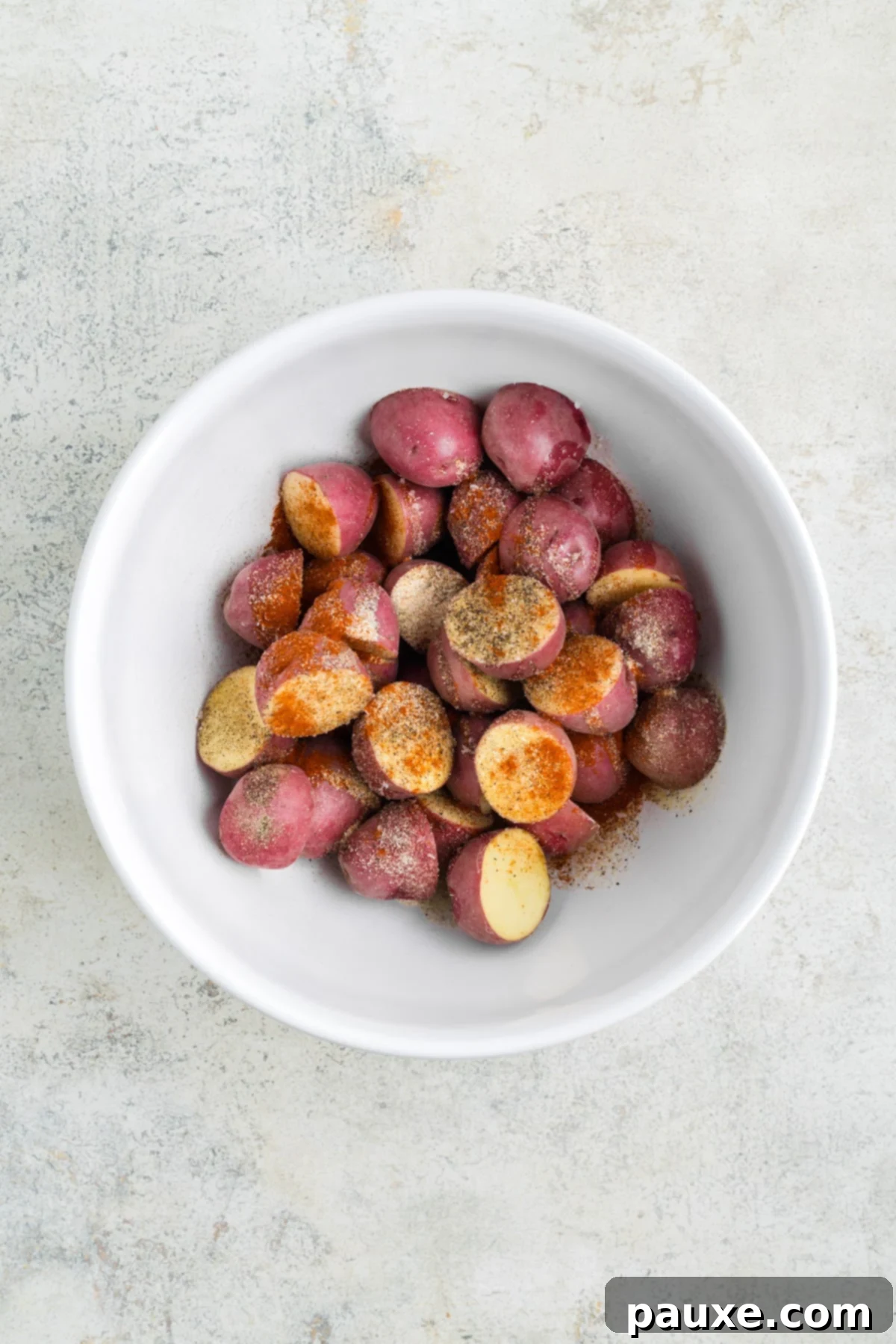A bowl of vibrant red potatoes generously topped with olive oil and a medley of seasonings, ready for tossing.